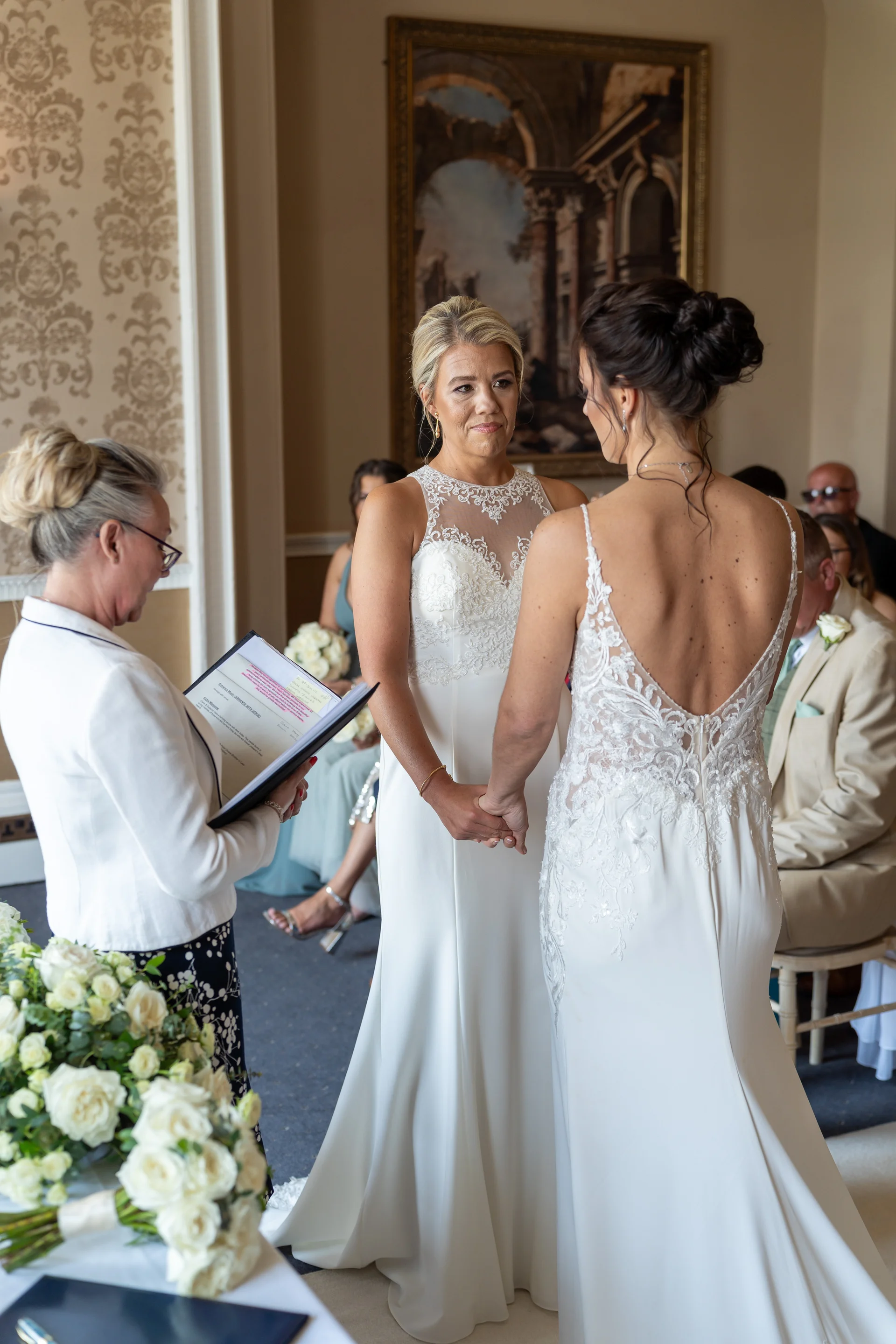 Two brides hold hands and exchange vows during their wedding ceremony inside a light filled venue, officiated by a celebrant.