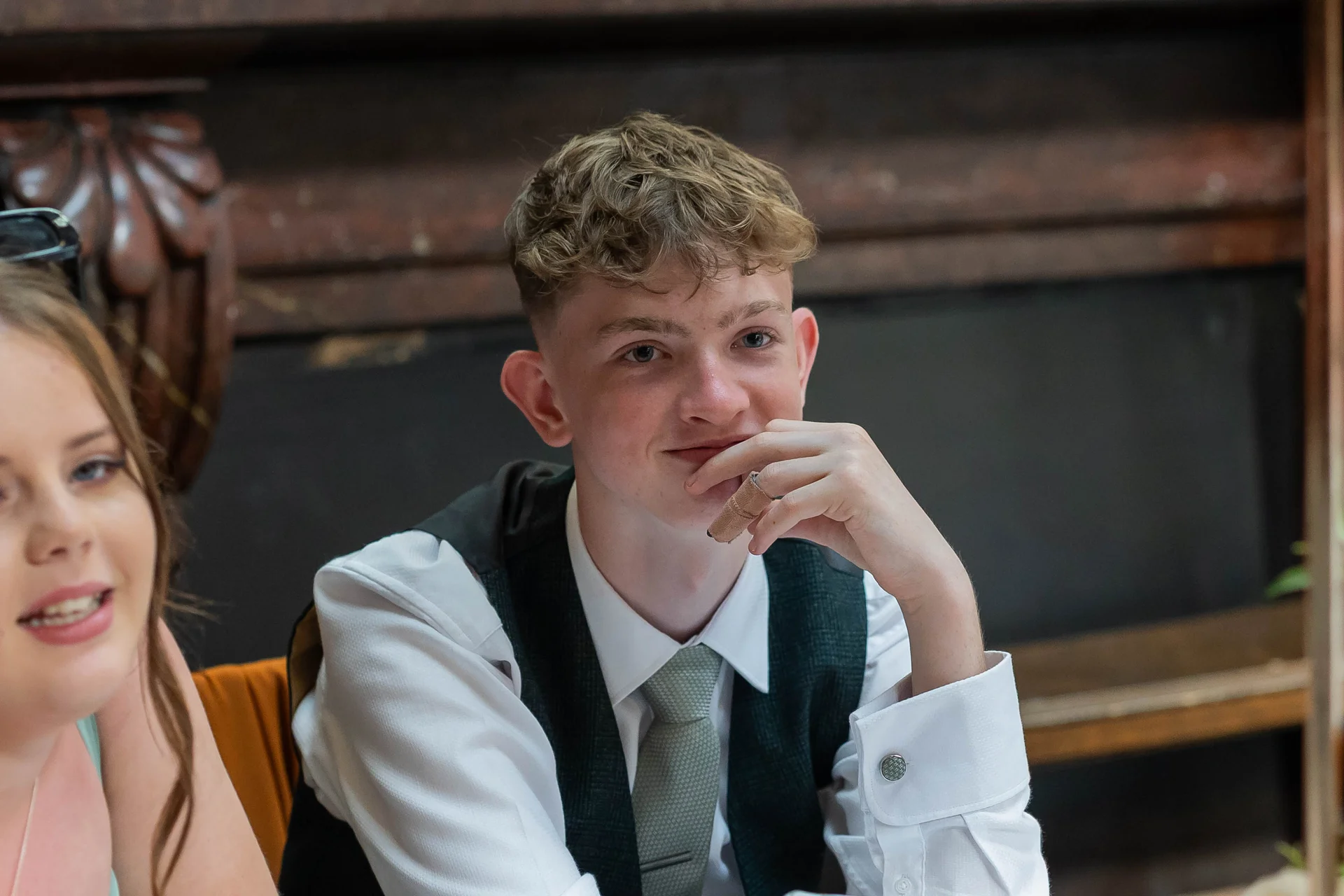 A young wedding guest in a waistcoat and tie sits smiling during the wedding reception speeches, with his hand resting near his chin.