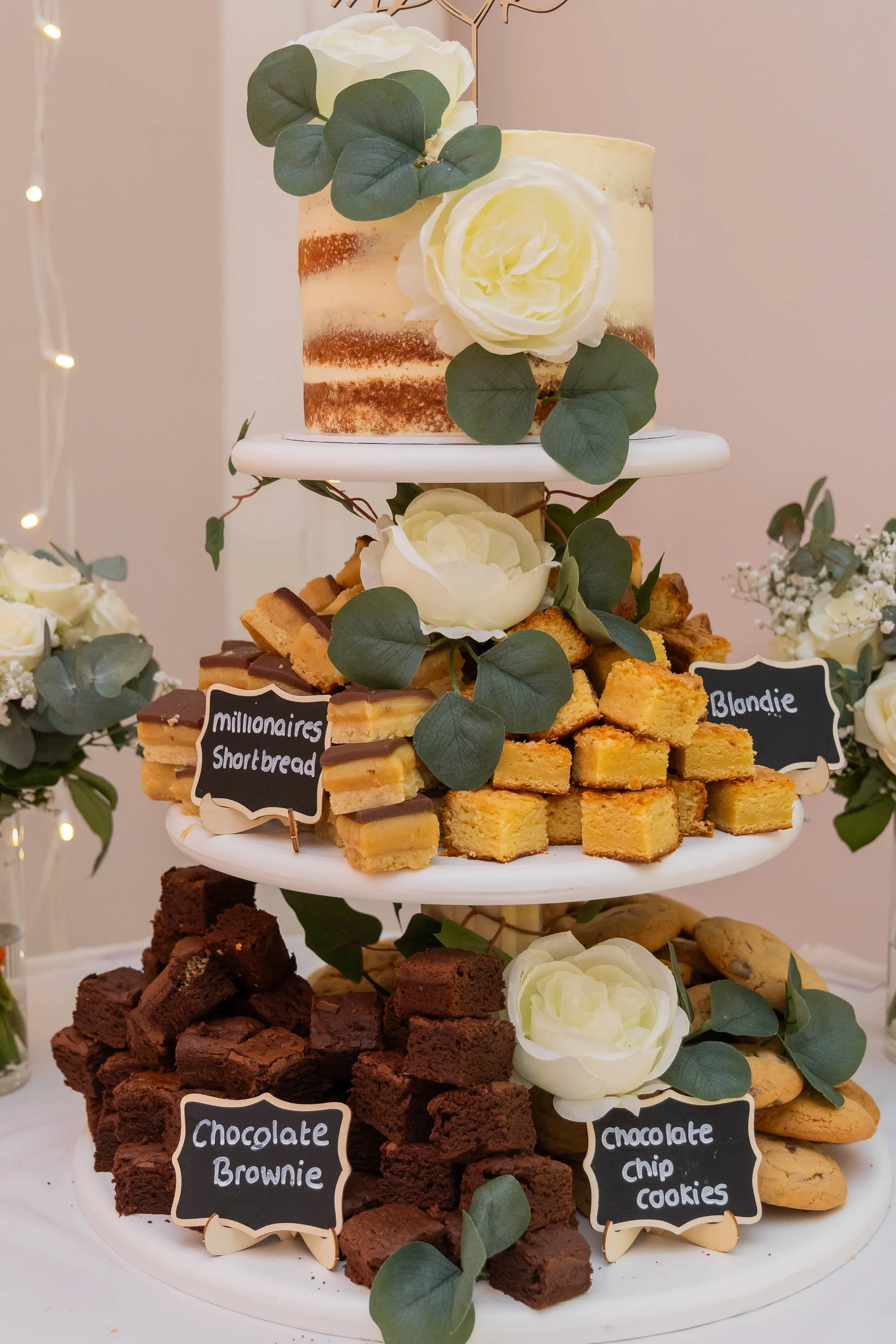 A magnificent three-tiered dessert display tower holds a semi-naked wedding cake, brownies, millionaire's shortbread, blondies, and cookies, beautifully adorned with white roses and eucalyptus leaves.