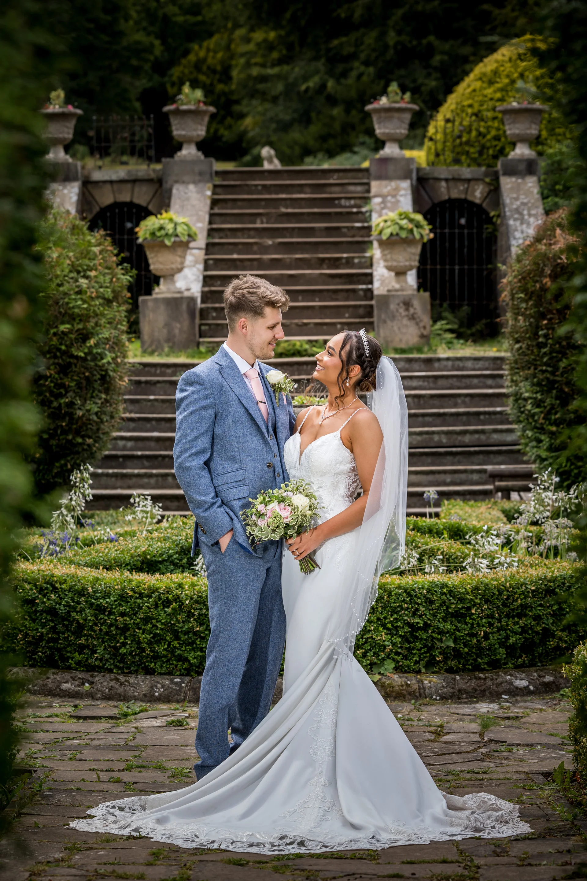 A newlywed couple stands smiling at each other in a manicured country house garden, positioned in front of an impressive stone staircase with urns.