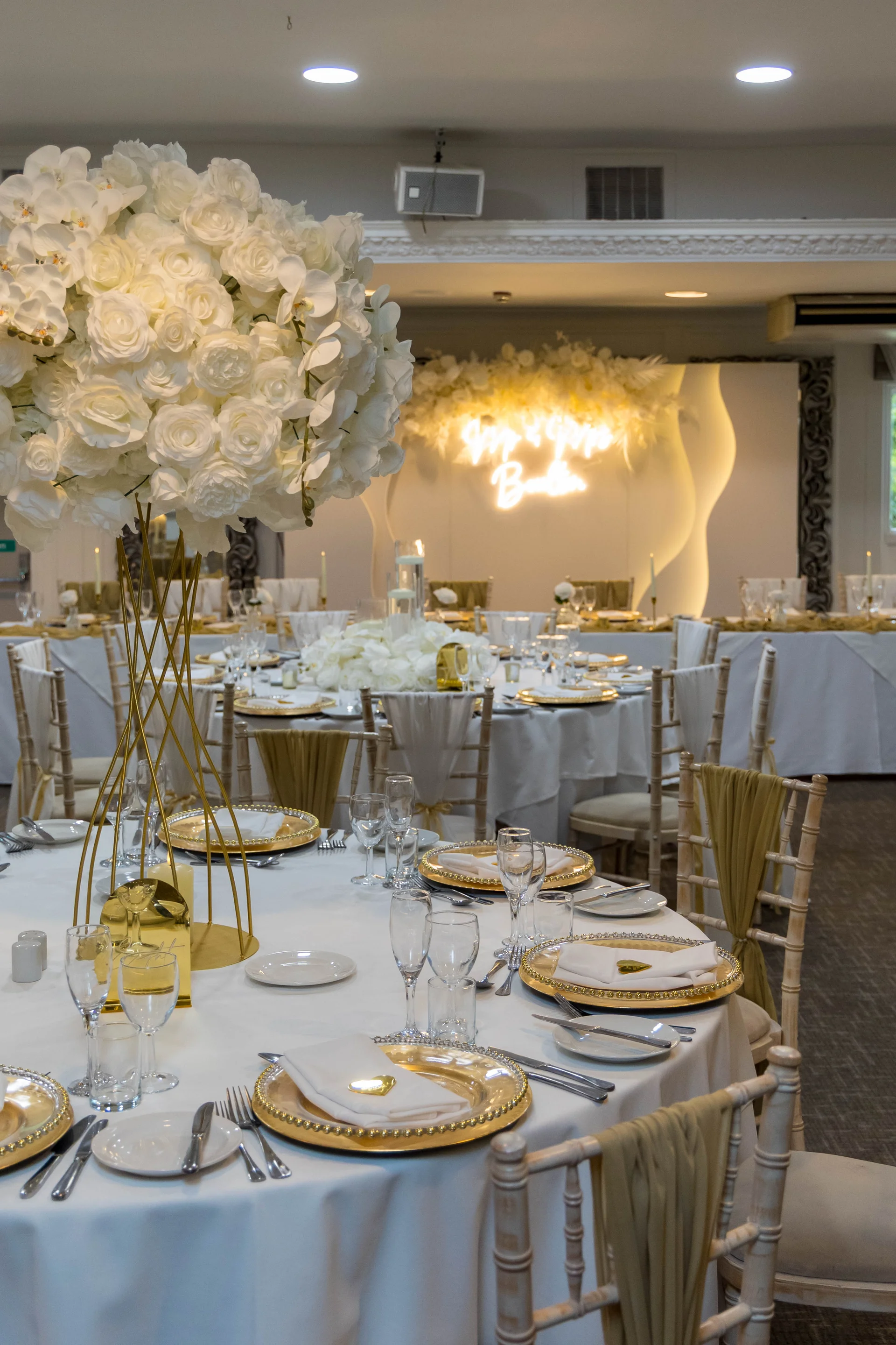 An elegant wedding reception tablescape features white floral centrepieces on tall gold stands, gold-rimmed charger plates, white linens, and a glowing neon backdrop.
