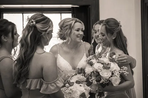 A joyful bride in a white lace gown shares a tender, laughing moment with her bridesmaids holding bouquets near a window in a black and white photograph.