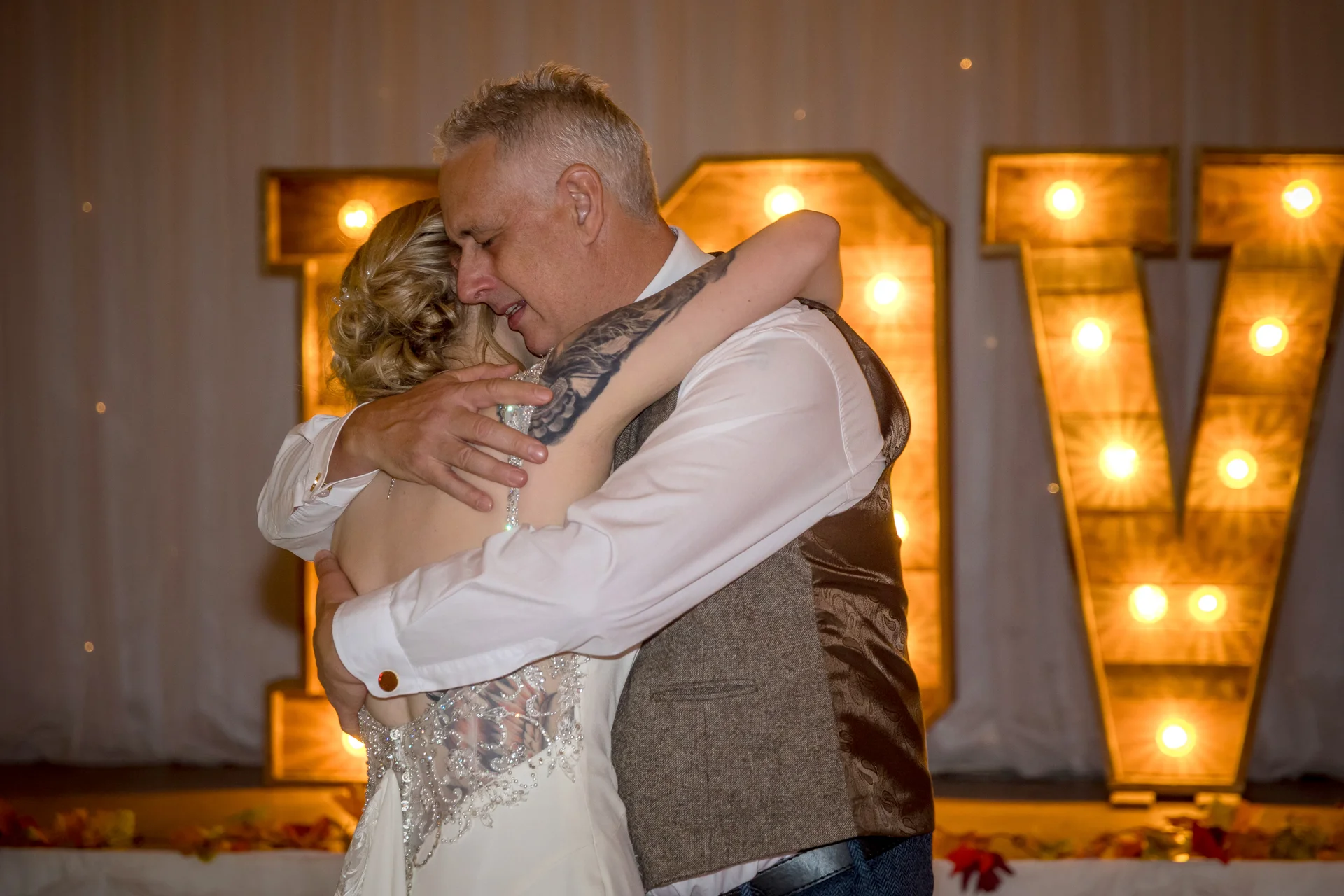 An emotional mature bride and groom share a tender embrace in front of illuminated marquee letters during their wedding reception.