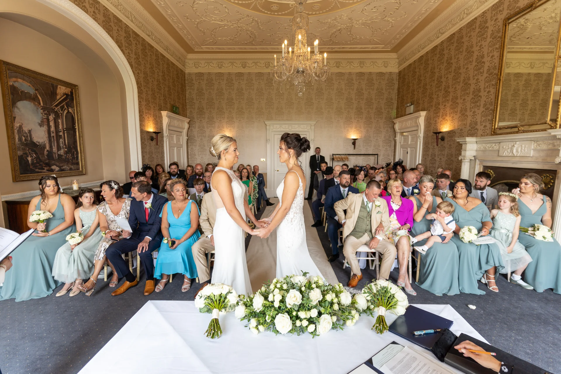 Two brides stand hand in hand exchanging vows at the altar of a grand stately home ceremony room filled with seated wedding guests.