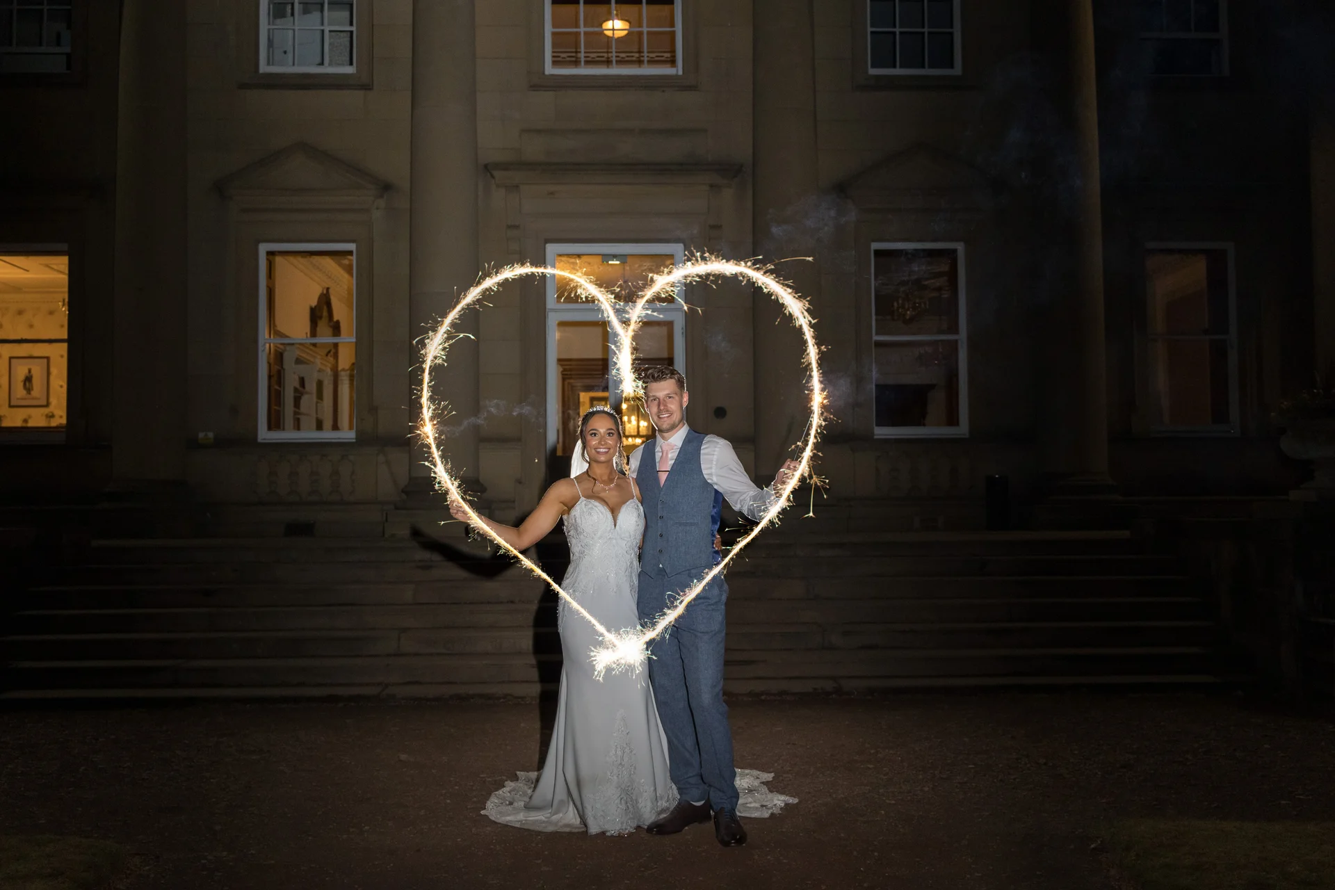 Newlyweds stand smiling outside a grand stately home at night, framed within a glowing light painting of a heart made using sparklers.