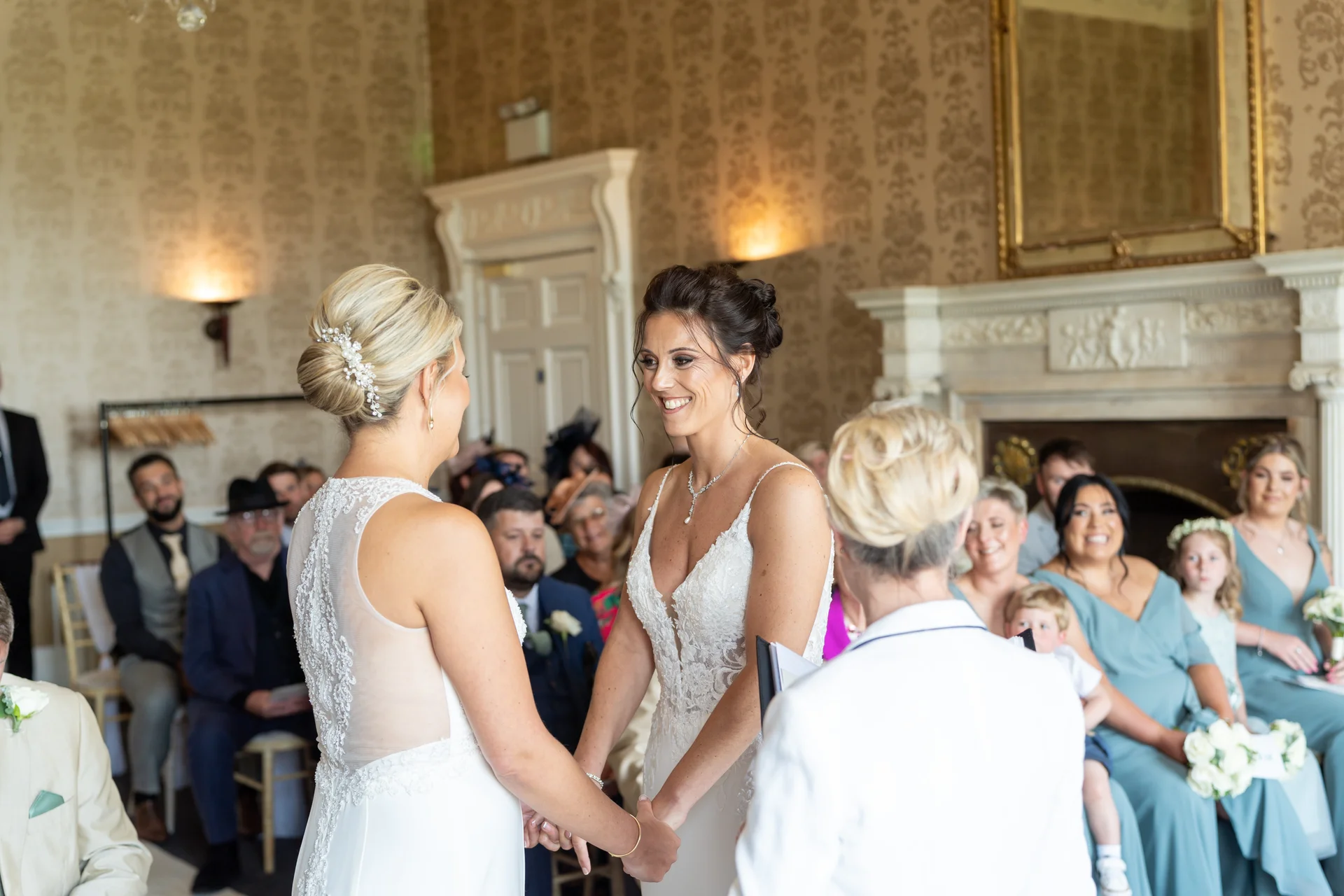 Two smiling brides hold hands facing each other during their indoor civil ceremony in an elegant room with a fireplace, surrounded by seated guests.