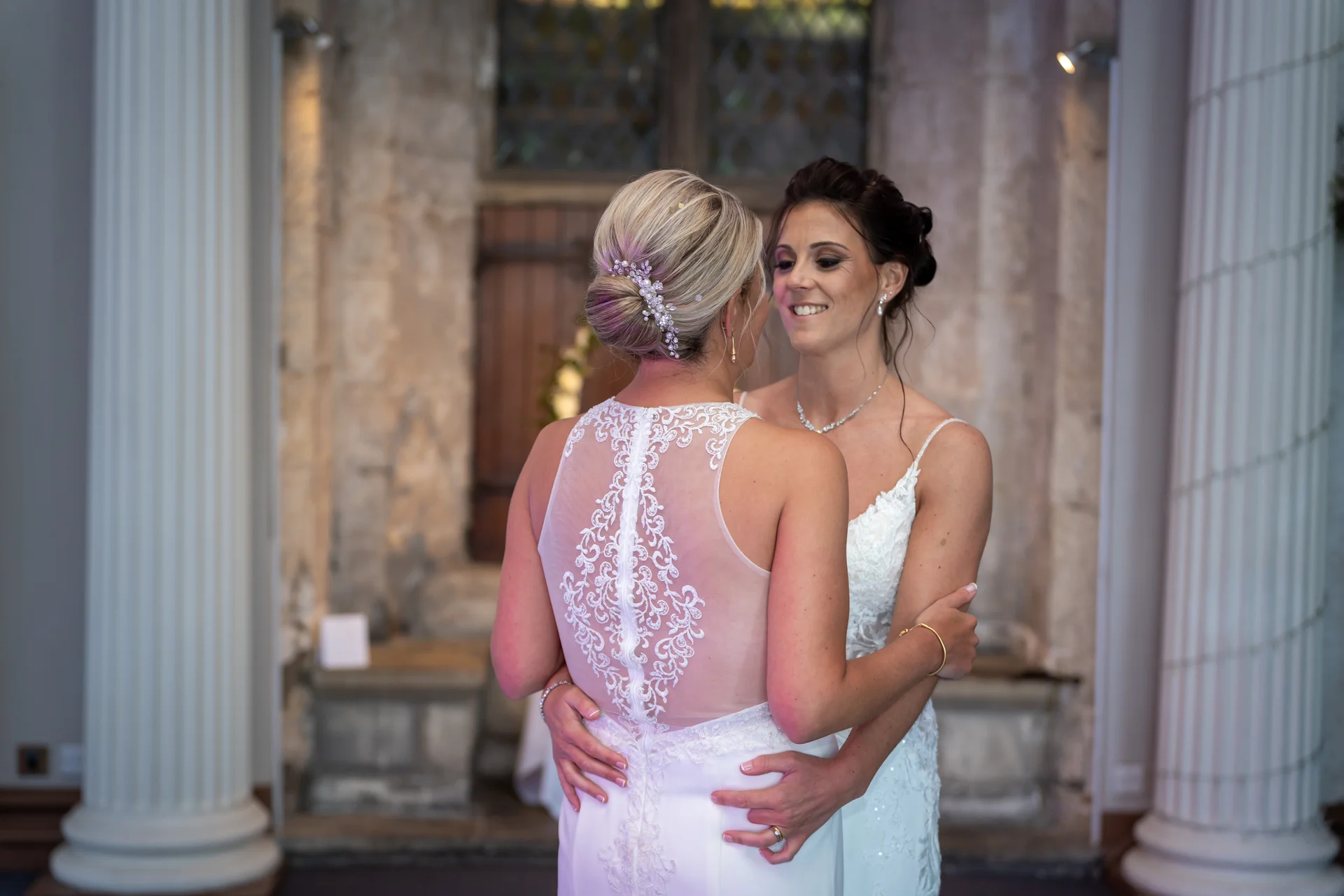 Two smiling brides embrace each other lovingly in front of a historic stone archway framed by white pillars.