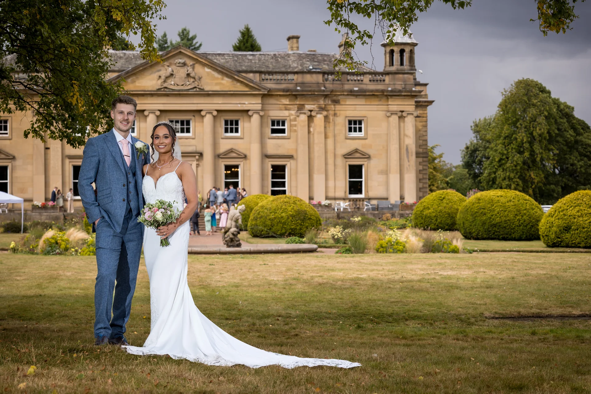 A newlywed couple poses together on the lawn in front of a grand stone country house on their wedding day.