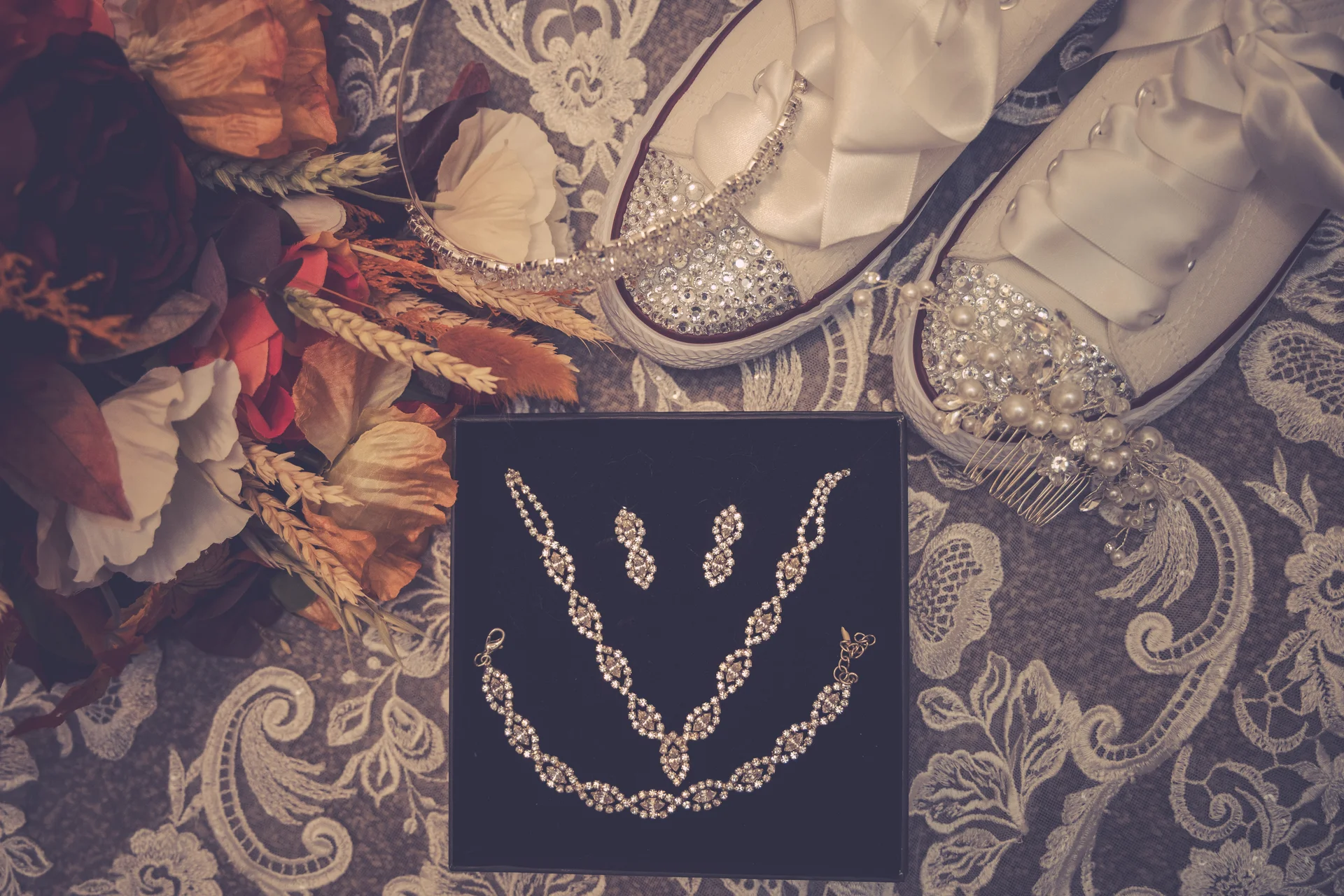 An artistic flat lay featuring sparkly embellished bridal trainers, a matching jewellery set in a black box, a crystal hair comb, and an autumnal bouquet resting on delicate lace fabric.