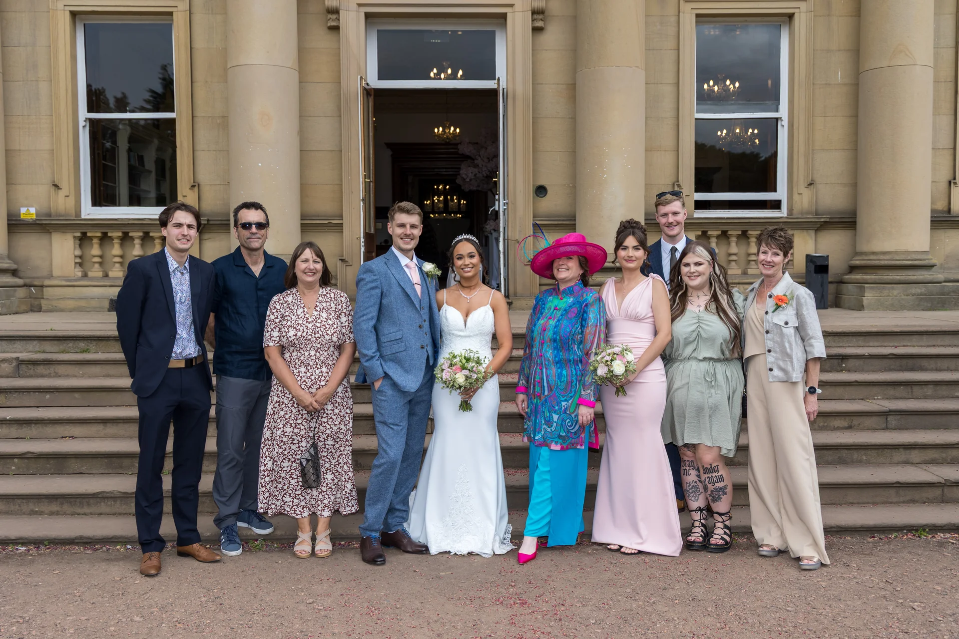 A bridal party and family members pose for a formal group photograph on the grand stone steps outside a stately manor house.
