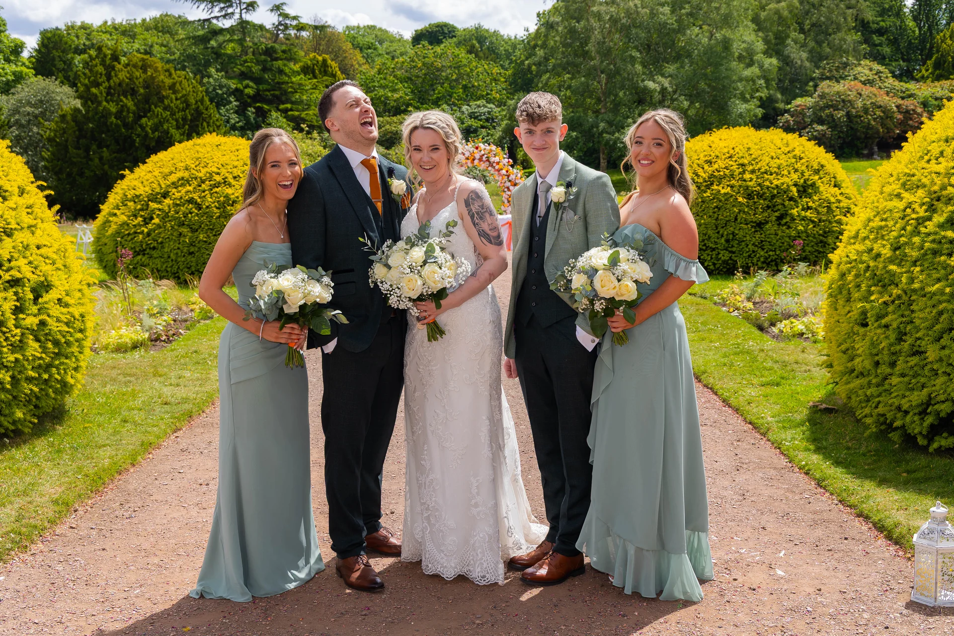 A joyful wedding party poses on a gravel garden path flanked by bright yellow shrubs, with the laughing groom holding a bouquet alongside the radiant bride and attendants.