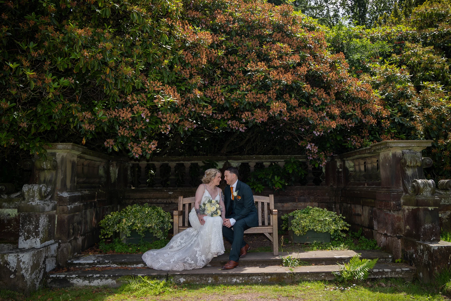 A newlywed couple smiling at each other while seated together on a wooden bench framed by an ornate stone balustrade and lush green foliage.