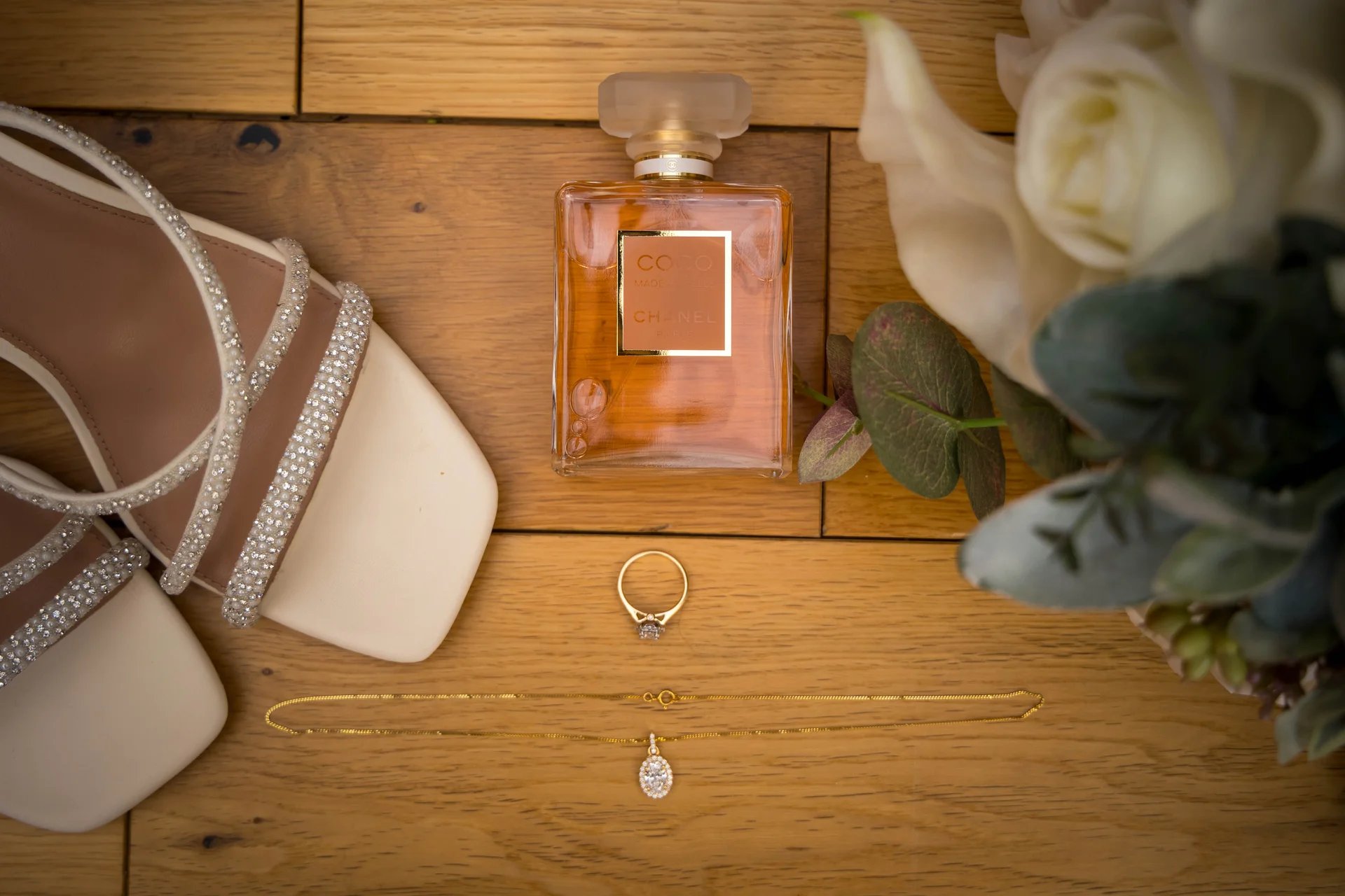 An elegant bridal flat lay featuring crystal-embellished strappy heels, a perfume bottle, a sparkling engagement ring, a delicate gold necklace, and white roses arranged on a wooden floor.