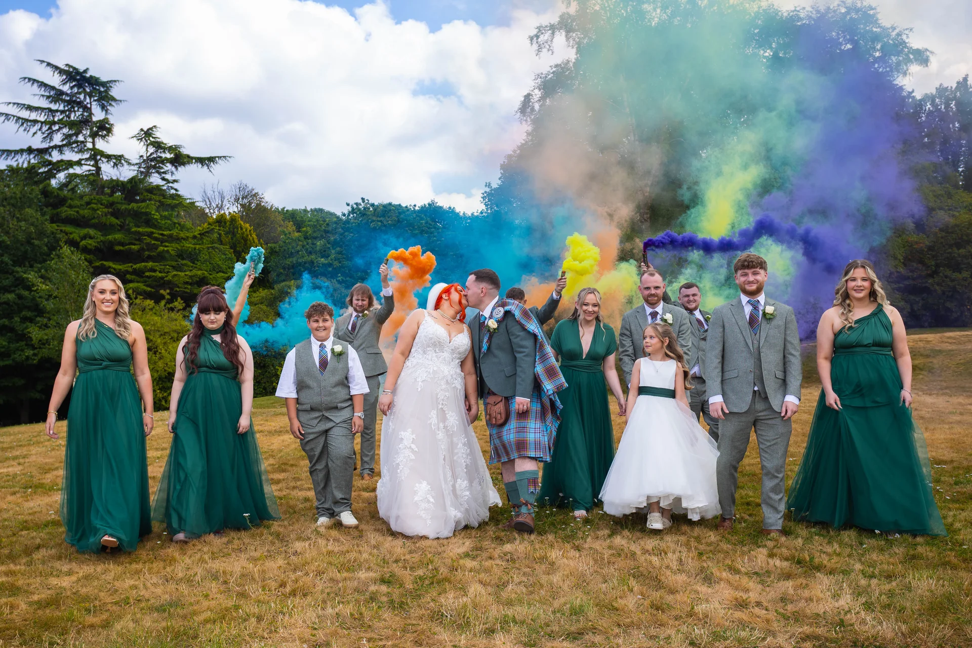 A bride and groom kiss outdoors surrounded by their wedding party holding colourful smoke grenades that fill the sky with vibrant hues.