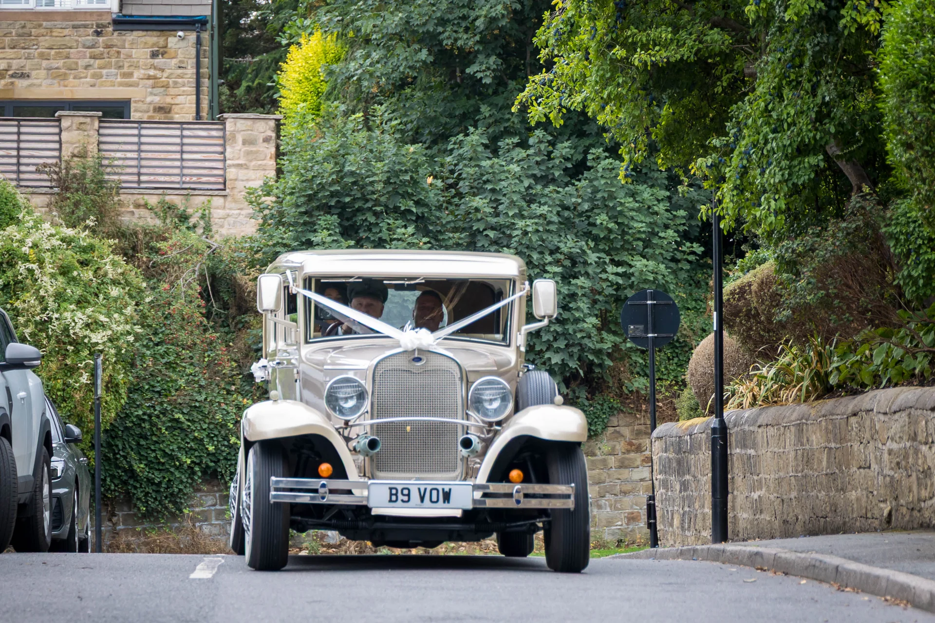 A vintage cream wedding car decorated with white ribbon drives along a country road bordered by stone walls and lush greenery.