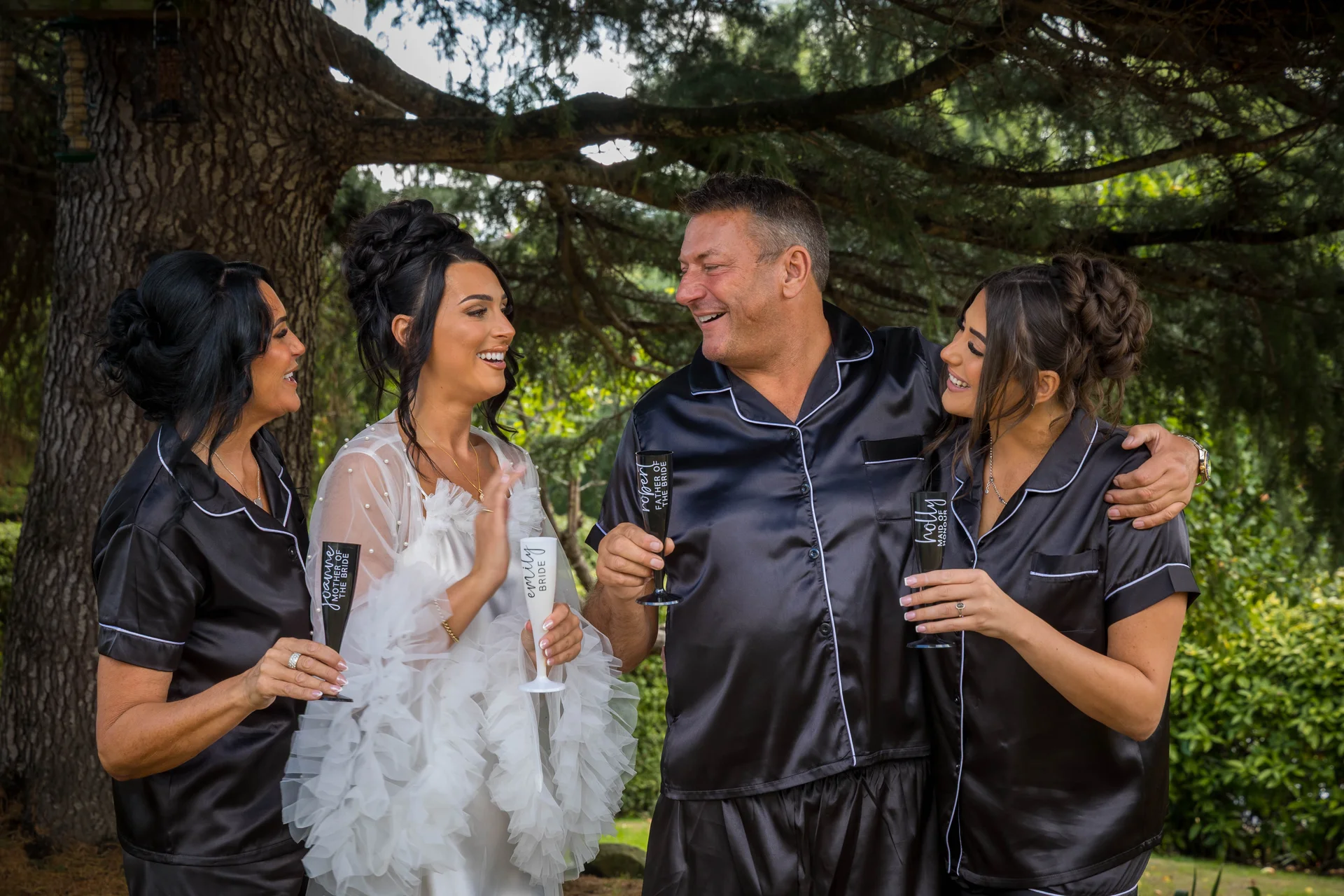 The smiling bride stands outdoors with her mother, father, and maid of honour, all wearing matching black satin pyjamas and holding custom champagne flutes.