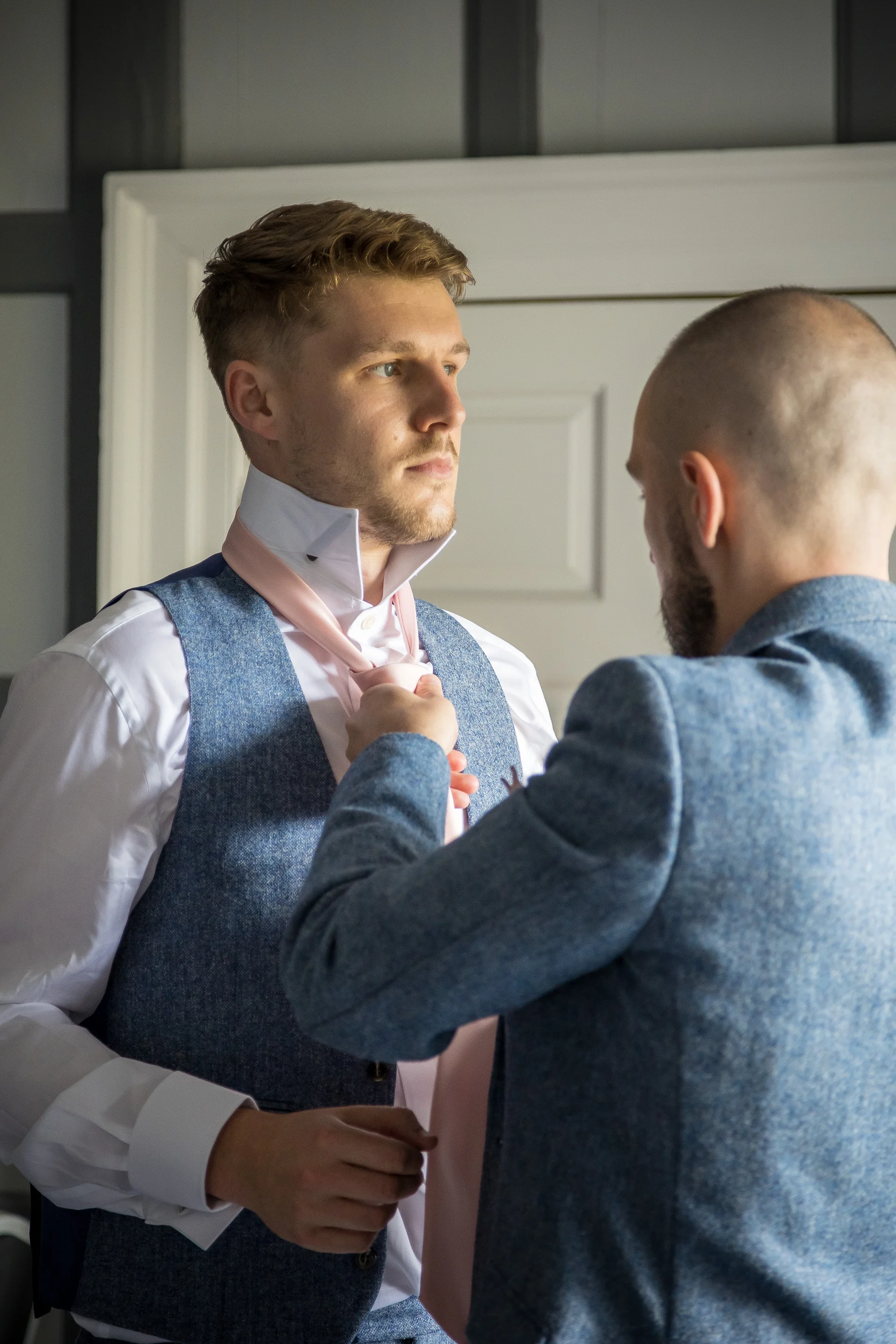 A groom wearing a blue tweed waistcoat looks thoughtfully ahead while another man adjusts his pink tie during morning preparations.