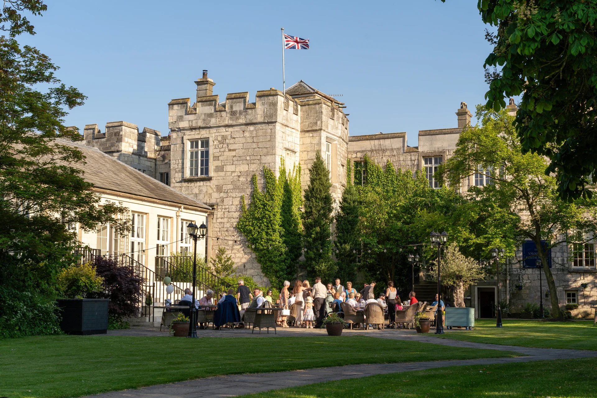 Wedding guests mingle during a sunny outdoor drinks reception in the courtyard of a historic stone castle venue.