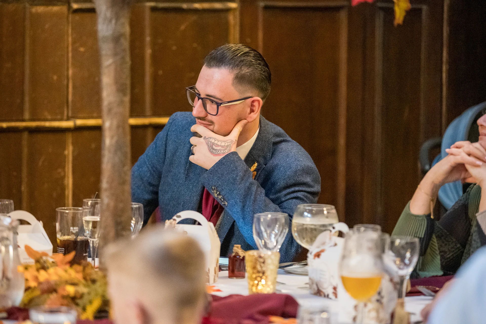 A wedding guest wearing a blue tweed suit and glasses rests his chin in his hand during the wedding breakfast.
