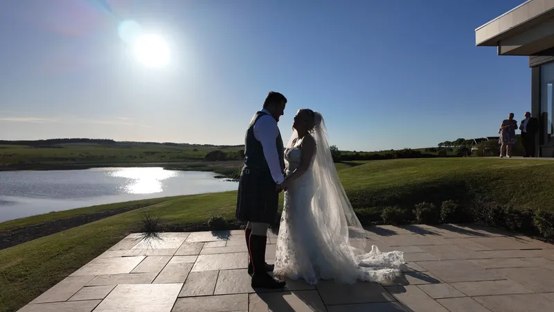 A newlywed couple stand on a stone patio overlooking a tranquil lake during golden hour, with the bride wearing a lace gown and long veil and the groom dressed in a traditional kilt.