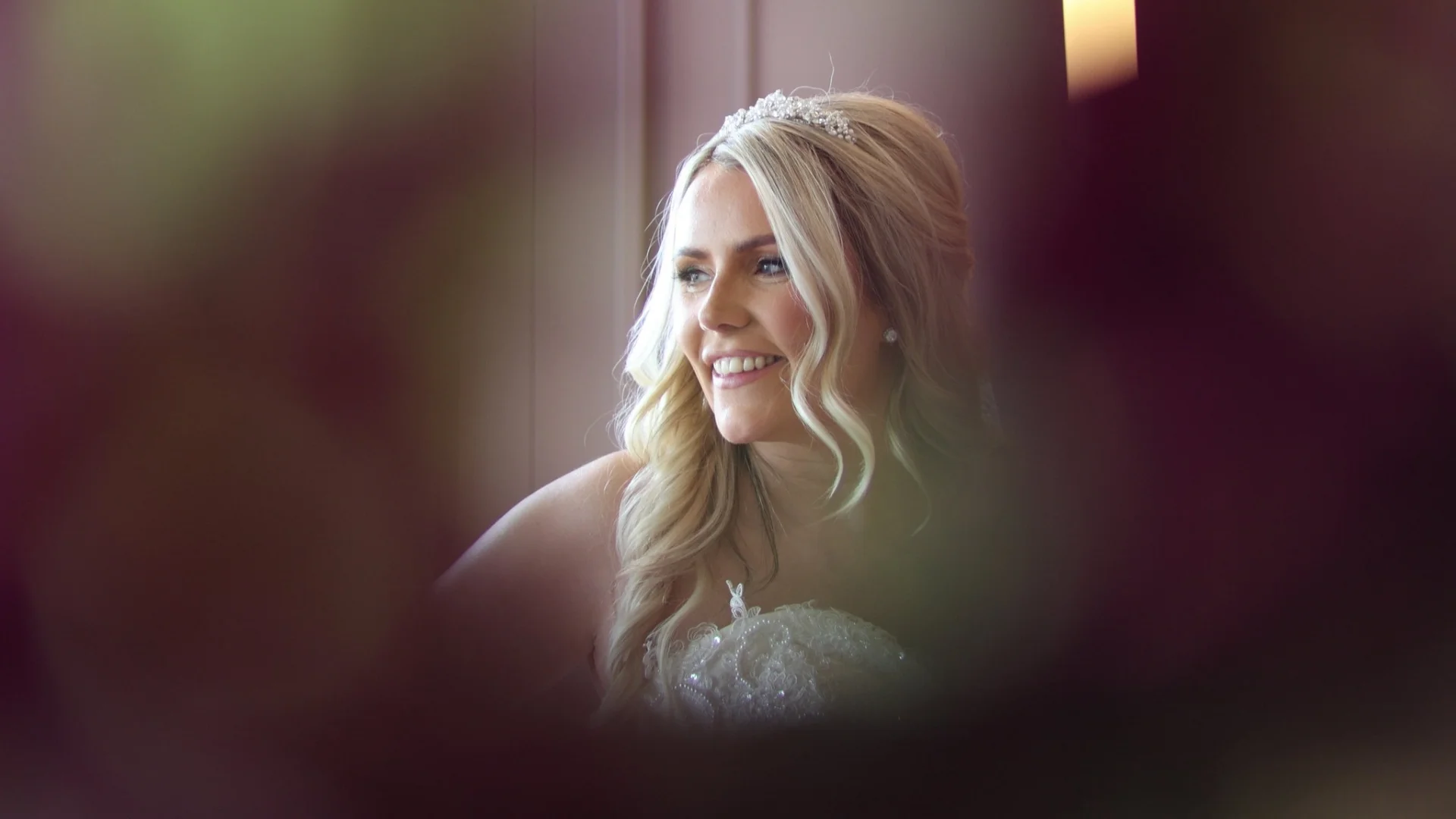 A smiling blonde bride wearing a crystal tiara and lace wedding dress is captured in a portrait framed by out-of-focus foliage.