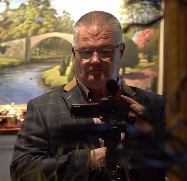 A man wearing glasses and a checked suit jacket stands in front of a landscape painting, capturing a reflection shot with a camera.
