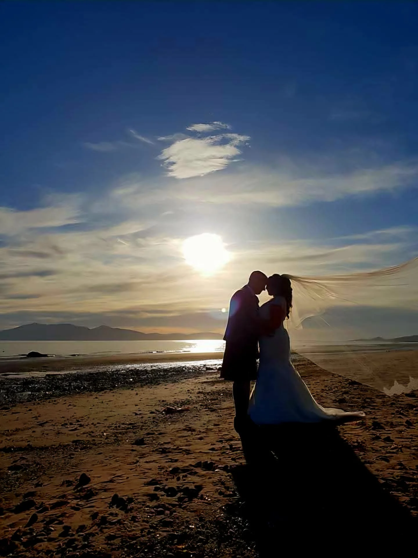 A newlywed couple shares a tender forehead kiss in silhouette on a sandy beach at sunset, with the bride's veil catching the wind against a brilliant dramatic sky.