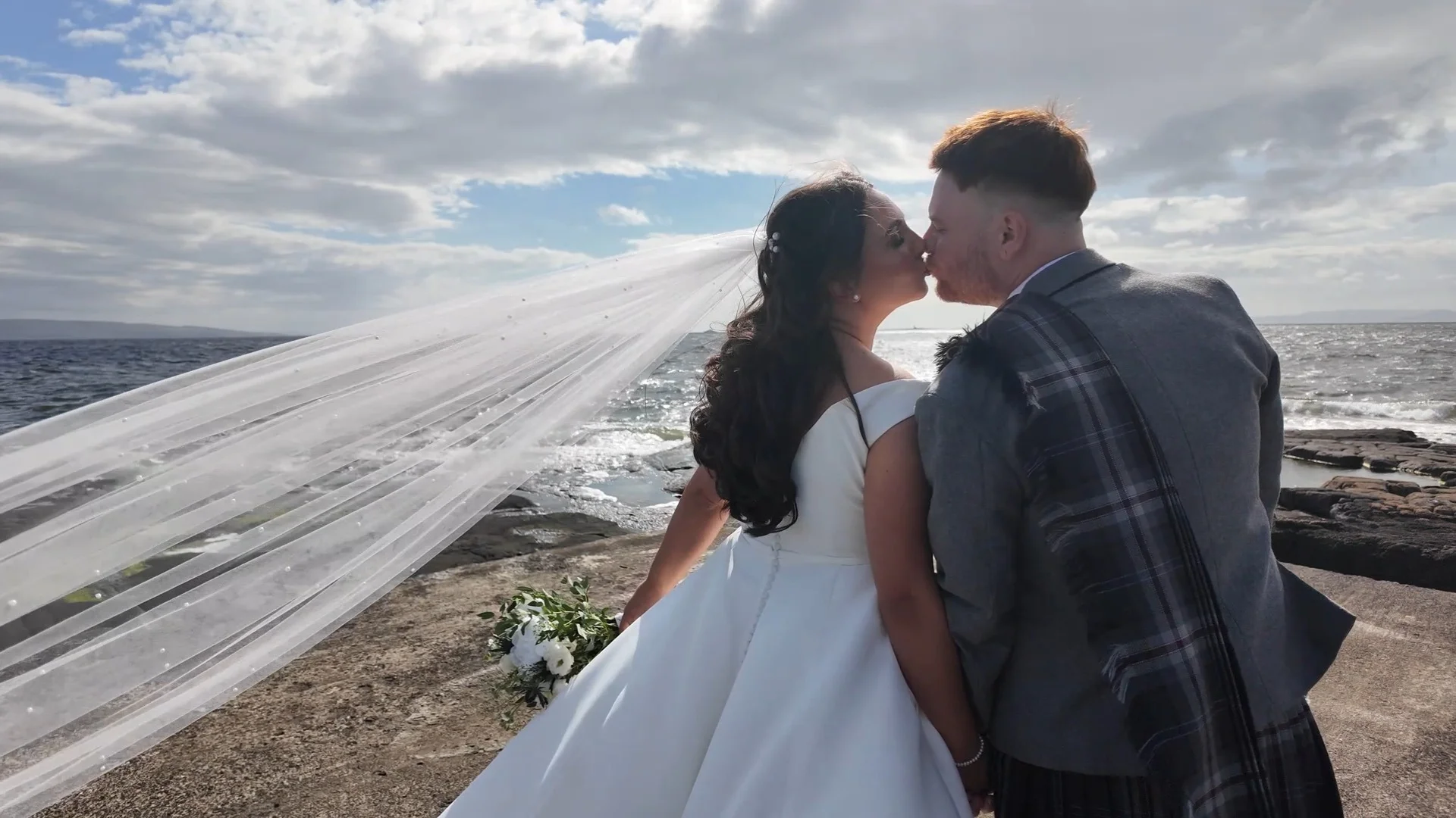 A newlywed couple shares a kiss on a coastal clifftop while the bride's long veil billows dramatically in the wind against a backdrop of the sea and cloudy sky.