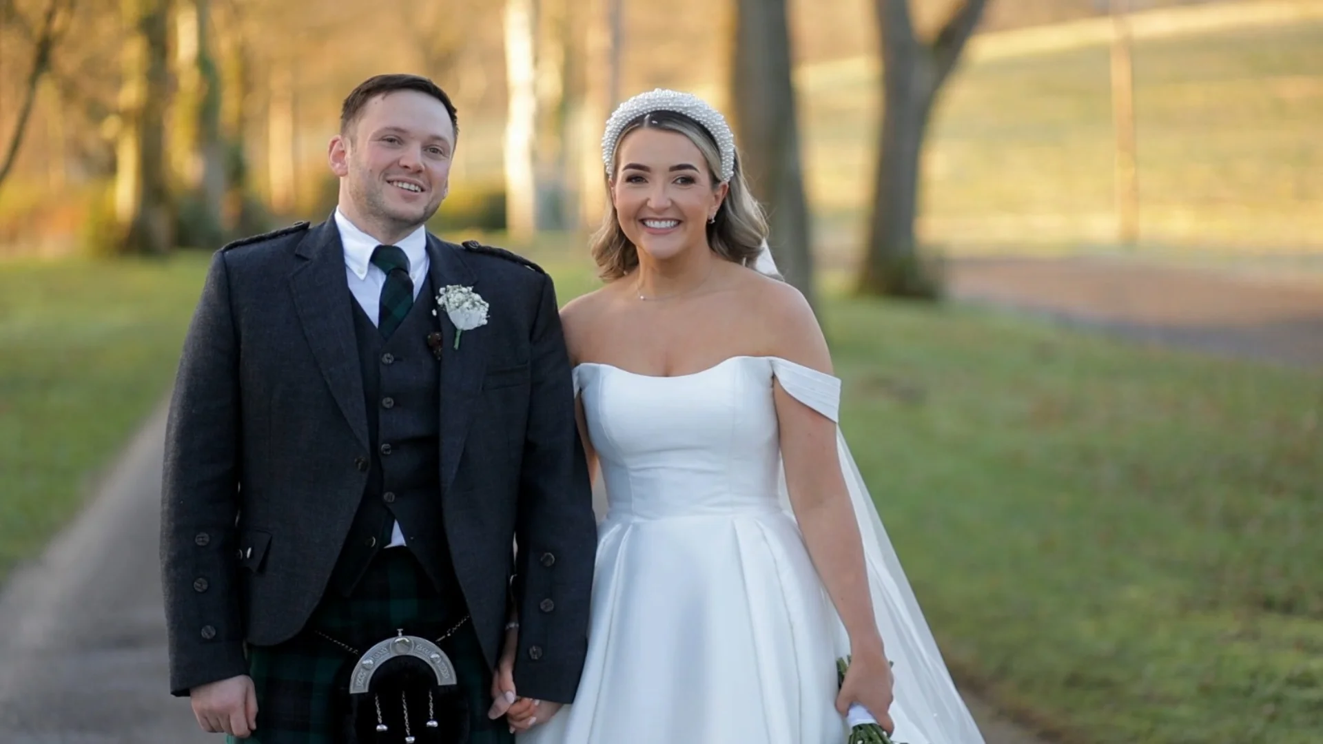A newlywed couple smiling together outdoors in a scenic park, with the groom wearing traditional Scottish highland dress and the bride in an off-the-shoulder white satin gown with a pearl headband.