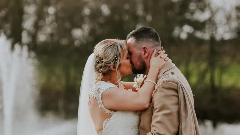A newlywed couple share a tender kiss outdoors by a fountain and woodland backdrop, with the bride wearing an intricate lace gown and veil.