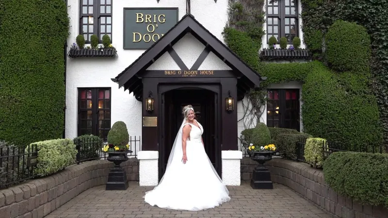 A smiling bride wearing a voluminous white ball gown and veil stands proudly in the entrance of the Brig O Doon House hotel.