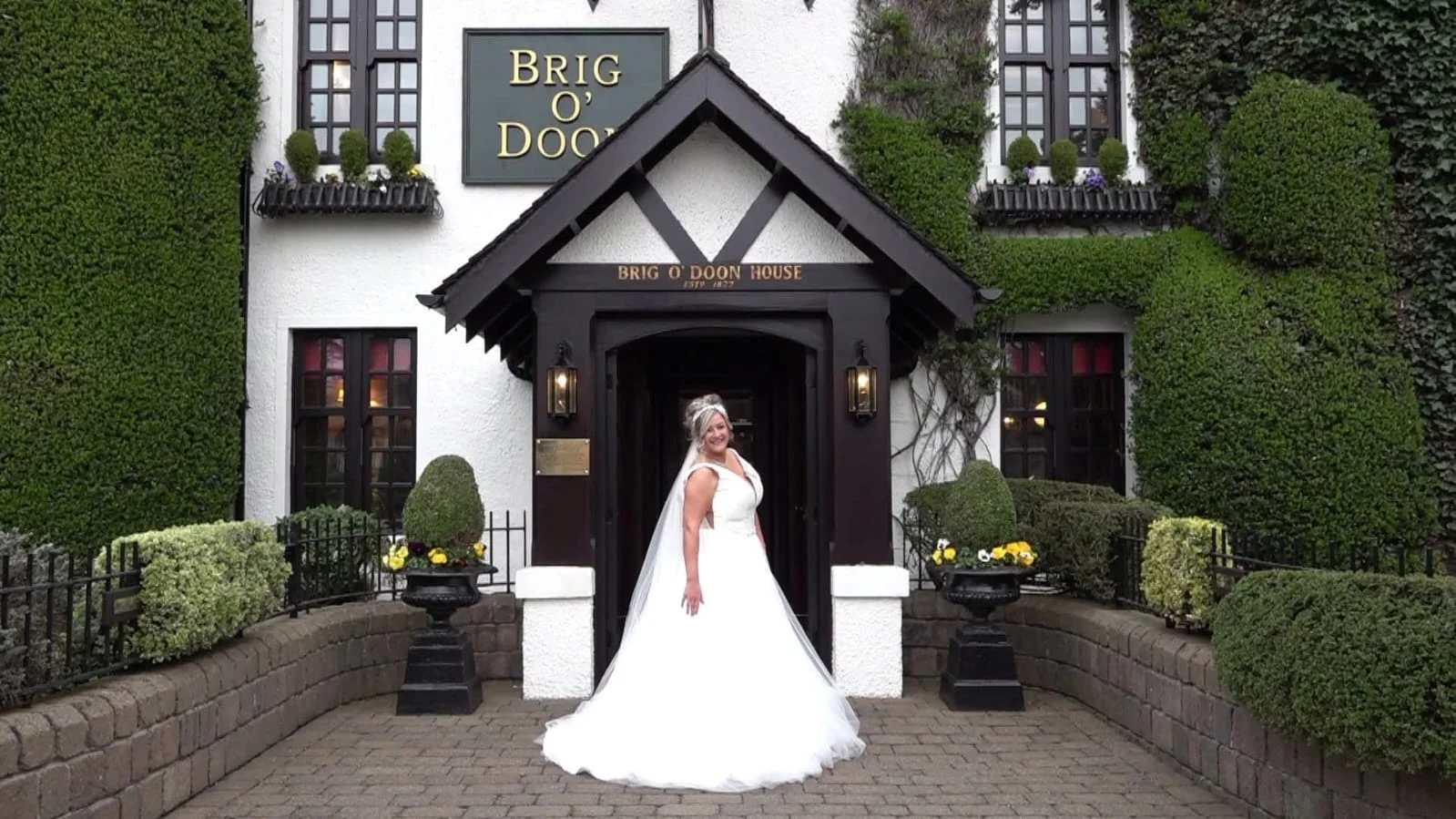 A smiling bride in a voluminous white a-line wedding gown and veil poses outdoors in the cobbled courtyard of the historic Brig O Doon House venue.