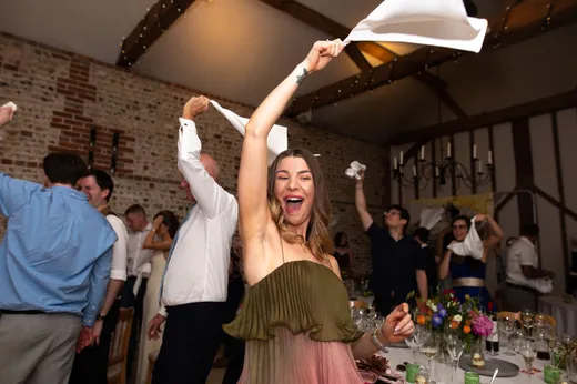 A joyful wedding guest waves a white napkin in the air during the wedding speeches inside a rustic stone barn reception venue.