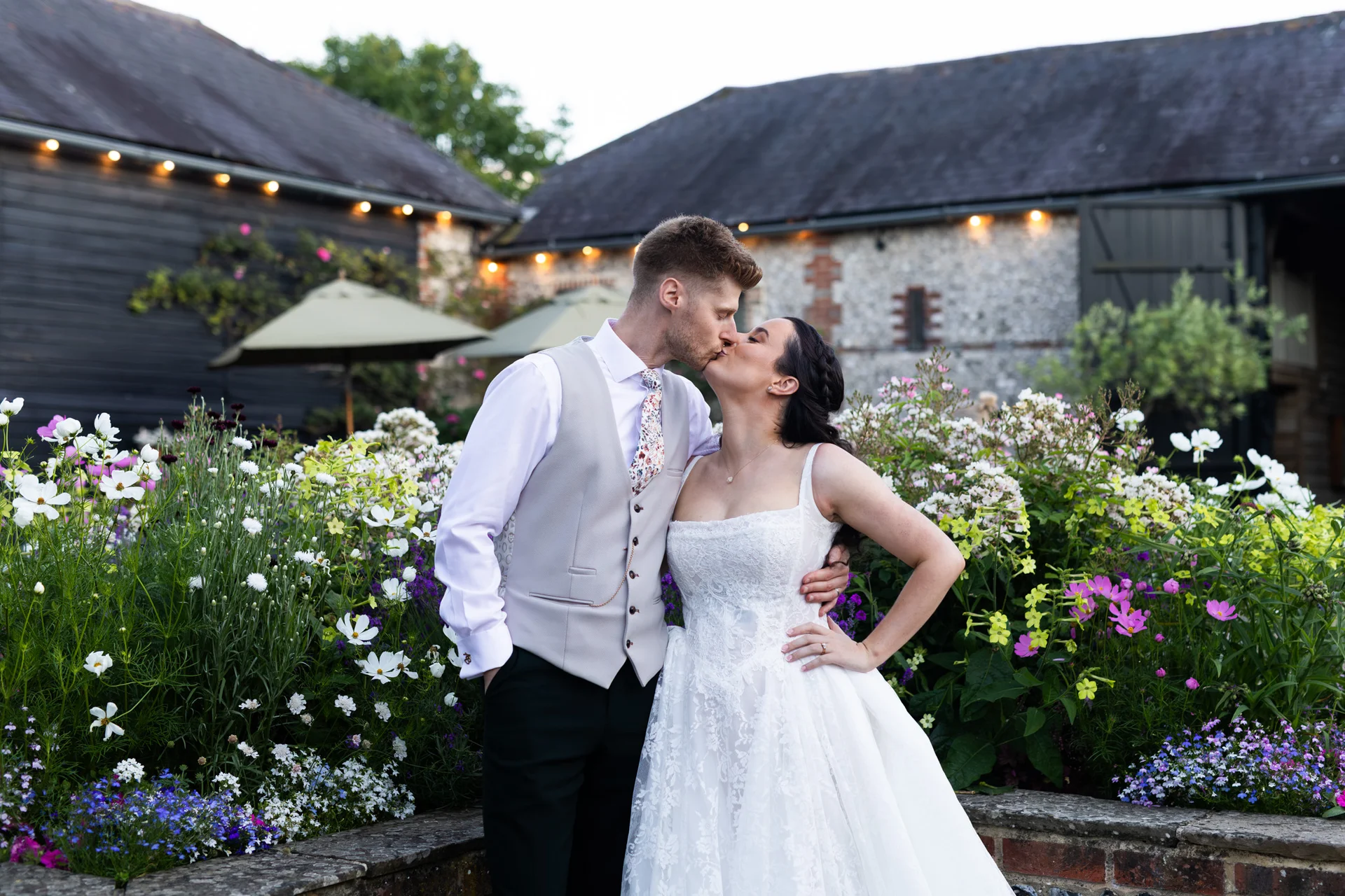 A newlywed couple share a tender kiss in front of a rustic stone barn surrounded by a vibrant wildflower garden during their summer wedding reception.