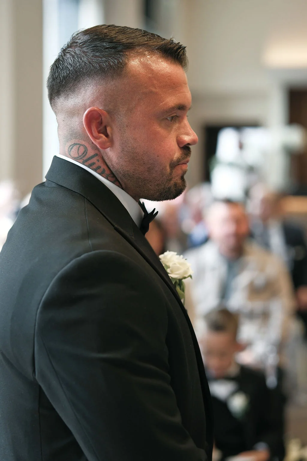 A close-up portrait of a groom wearing a black suit, bow tie, and white buttonhole as he waits at the altar during the ceremony.