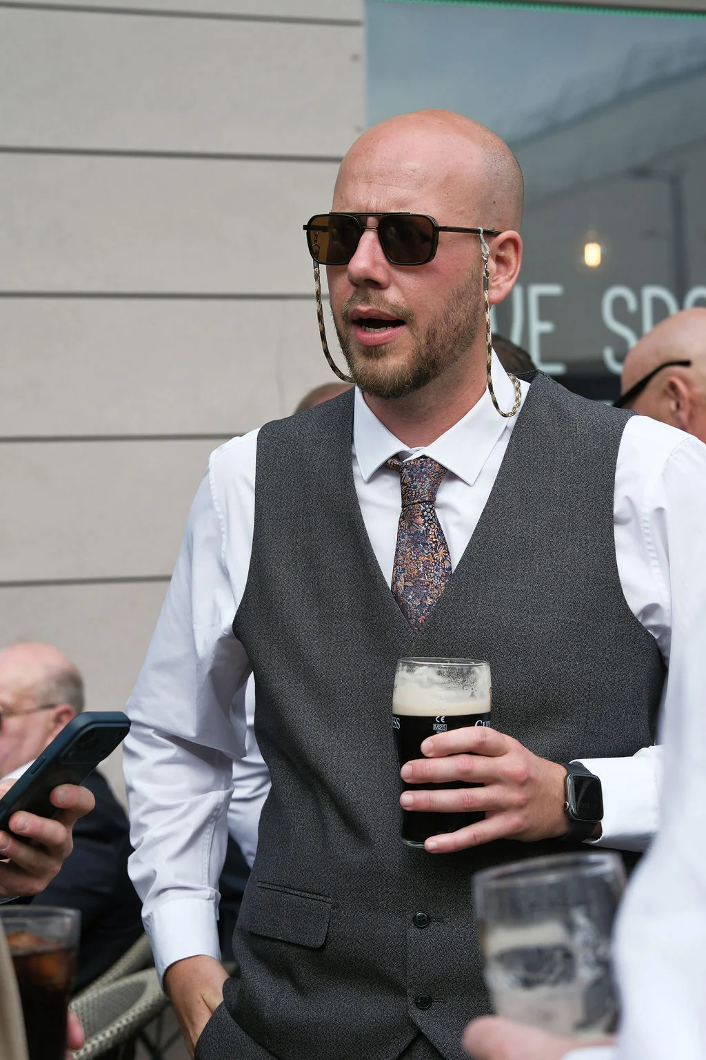 A bald wedding guest wearing sunglasses, a grey waistcoat, and a patterned tie holds a pint of stout during the drinks reception.