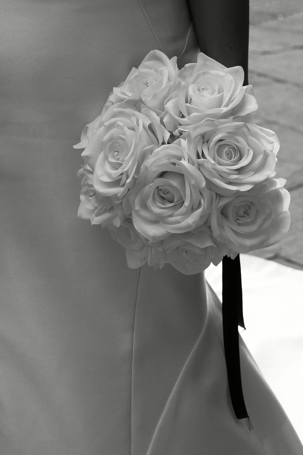 A close-up of a bride holding a round bouquet of white roses featuring subtle crystal centres against her smooth satin wedding gown.
