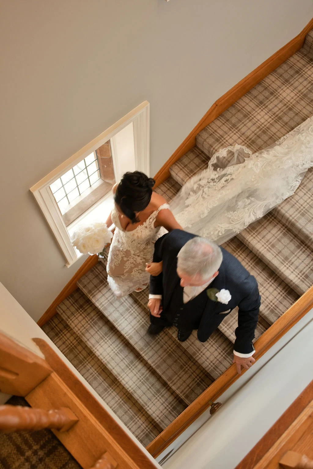 A high-angle shot captures a bride holding a white bouquet while walking down a tartan-carpeted staircase linked arm-in-arm with her father in a dark suit.
