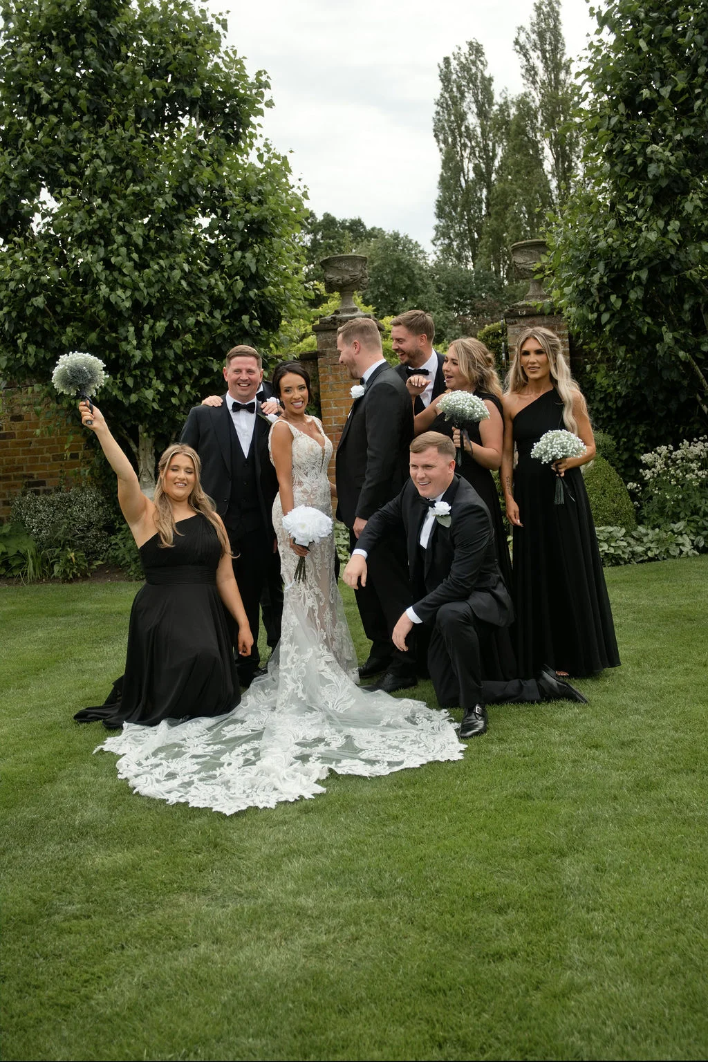 The bride and groom pose in a lush garden setting surrounded by groomsmen in black tuxedos and bridesmaids in matching black gowns holding gypsophila bouquets.