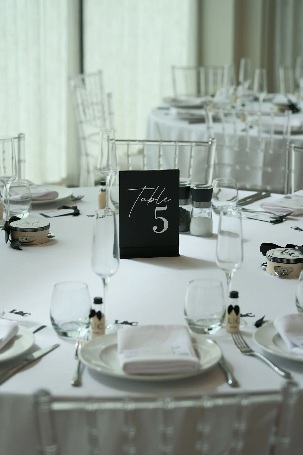 An elegantly styled round wedding table features a black table number card, white linens, crystal glassware, and monochrome favours in a sophisticated modern setting.