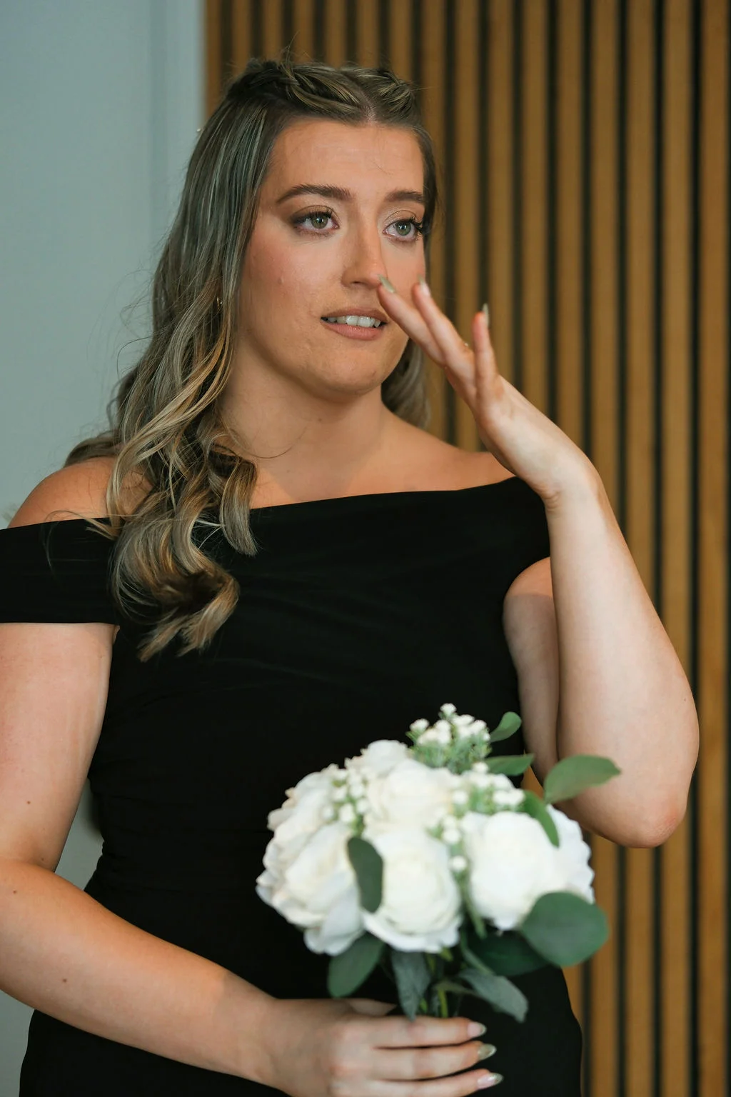 A bridesmaid in a black off-the-shoulder dress holding a hand-tied bouquet of white roses while wiping away an emotional tear.