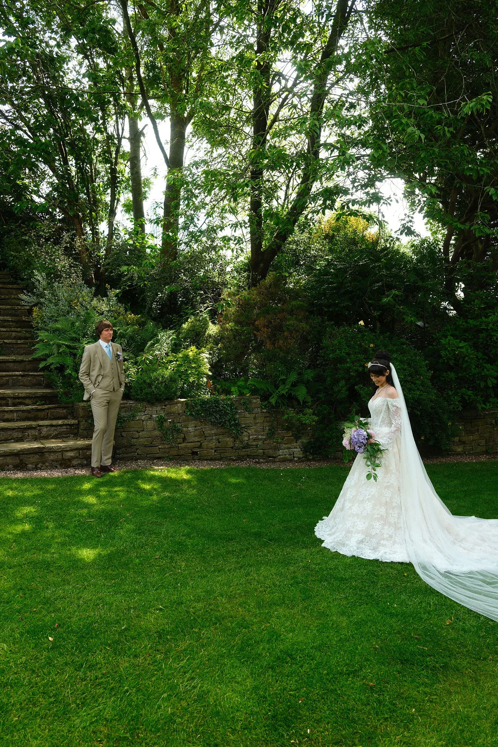A groom in a beige three-piece suit looks admiringly at his bride standing on a manicured lawn holding a cascading bouquet and wearing an off-the-shoulder lace gown with a long cathedral veil, framed by lush green trees and a stone staircase in a country garden setting.