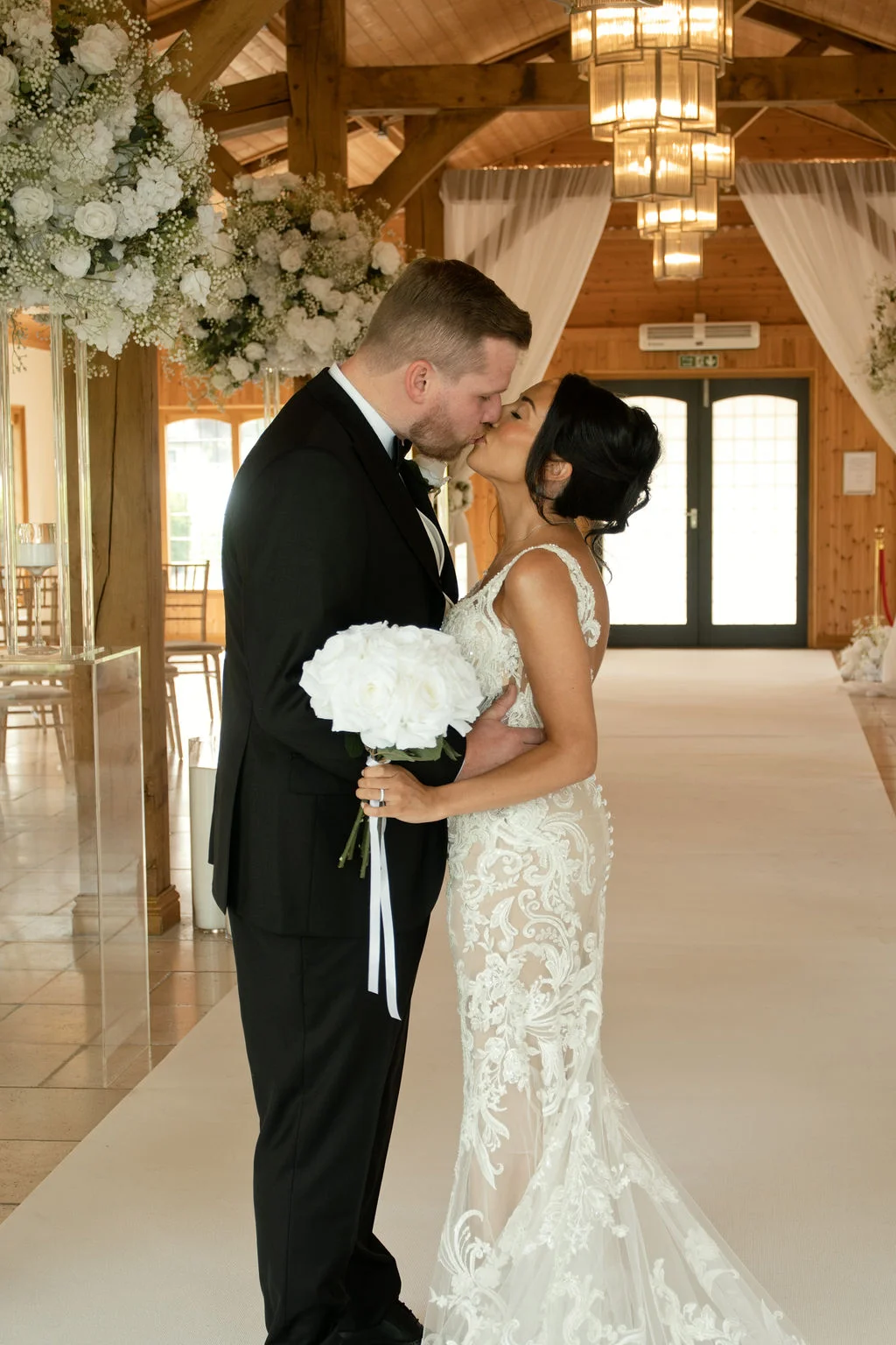 A newlywed couple shares their first kiss inside a rustic oak framed barn featuring elegant white floral arrangements and a vaulted wooden ceiling.
