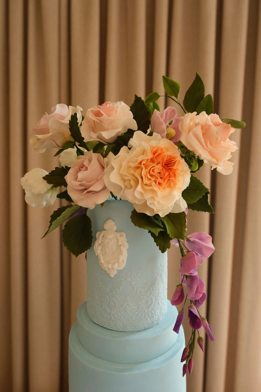An elegant pastel blue wedding cake features intricate lace detailing, a white cameo applique, and an elaborate topper of handcrafted sugar roses and trailing sweet peas.