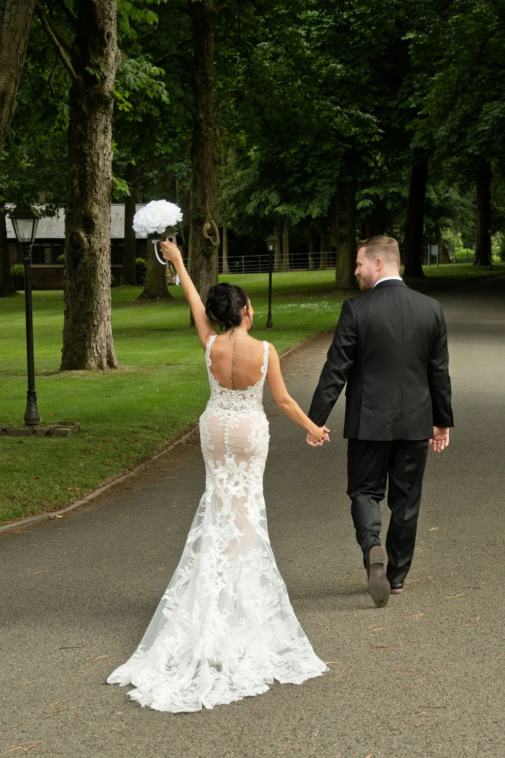 A newlywed couple walk hand in hand down a tree lined path, with the bride holding her white bouquet up in celebration.