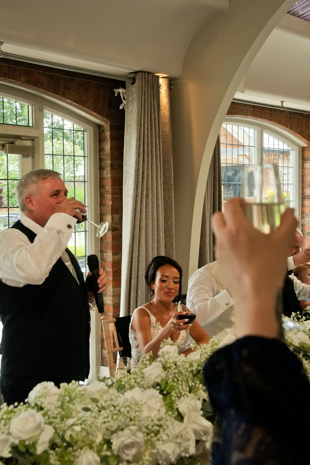 A guest raises a glass during the wedding speeches as family members at the top table share a toast surrounded by white floral arrangements.