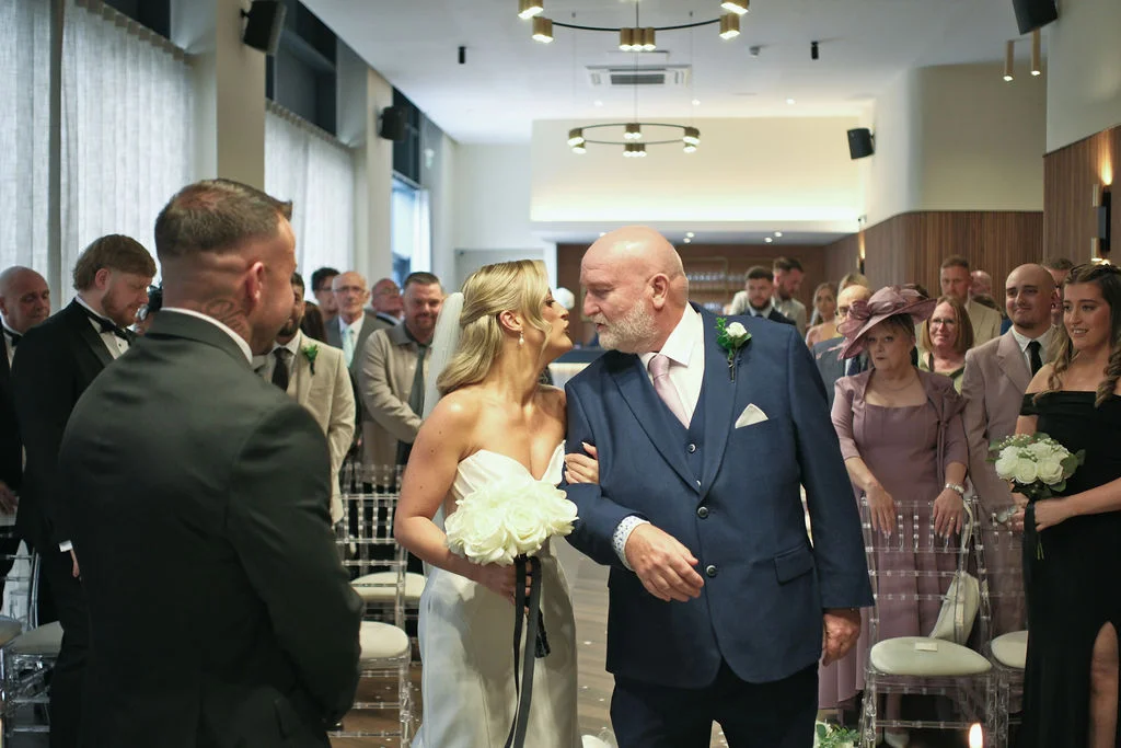 The bride walking down the aisle with her father, holding a white rose bouquet while approaching the groom at a hotel wedding ceremony.