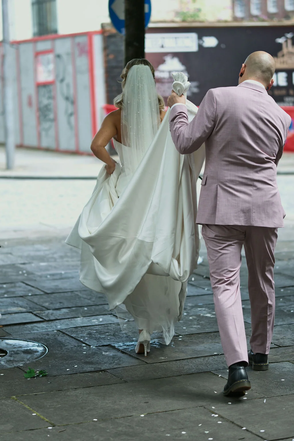 A newlywed couple walks away down a city street, with the bride holding up her white satin gown and wearing a pearl-embellished veil, while the groom wears a pink suit.