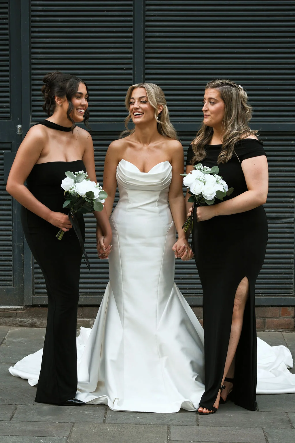 A smiling bride in a white satin strapless gown stands holding hands with two bridesmaids wearing black dresses and carrying white rose bouquets.