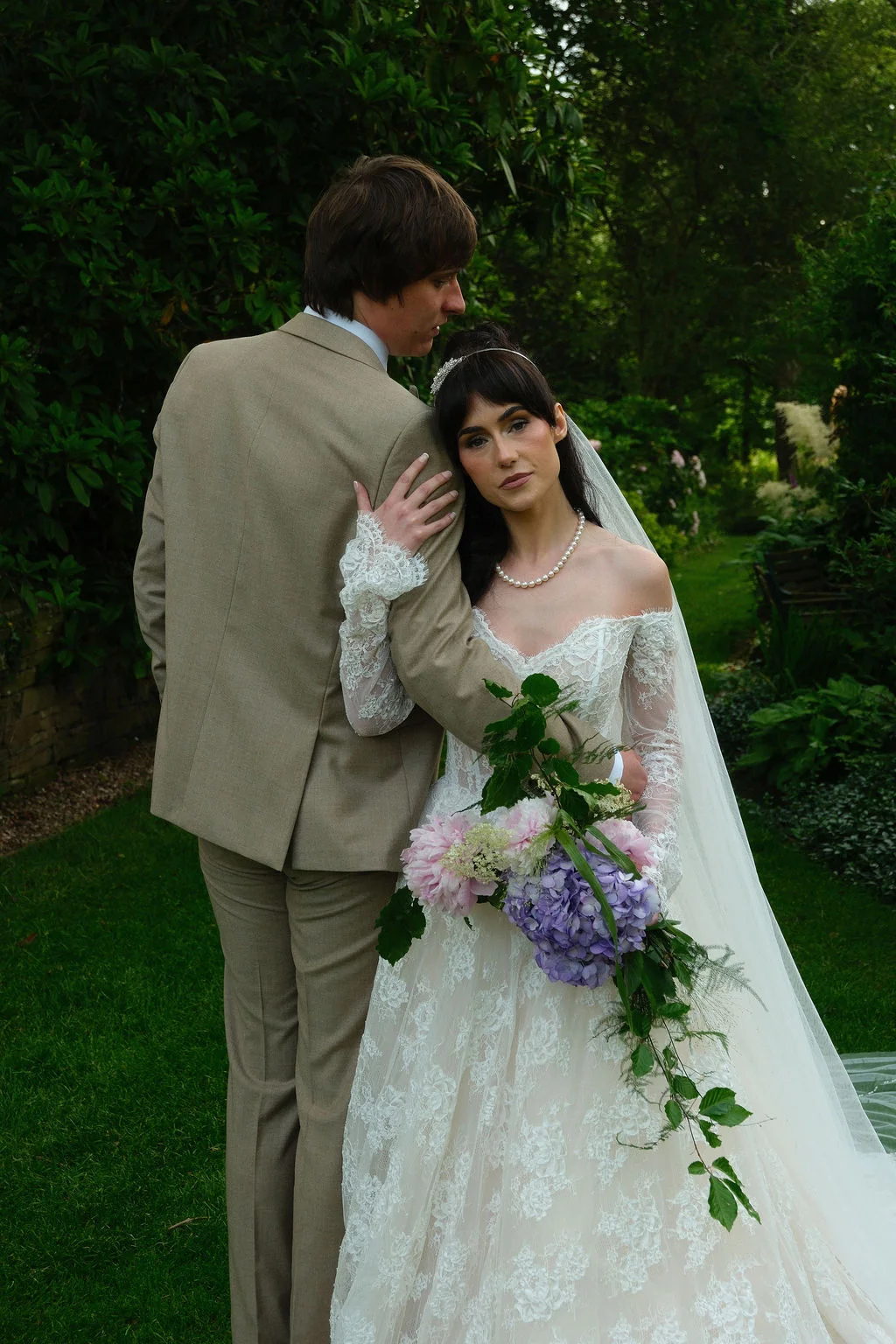 A bride with dark hair resting her head on the groom's shoulder while holding a cascading floral bouquet in a lush green garden setting.